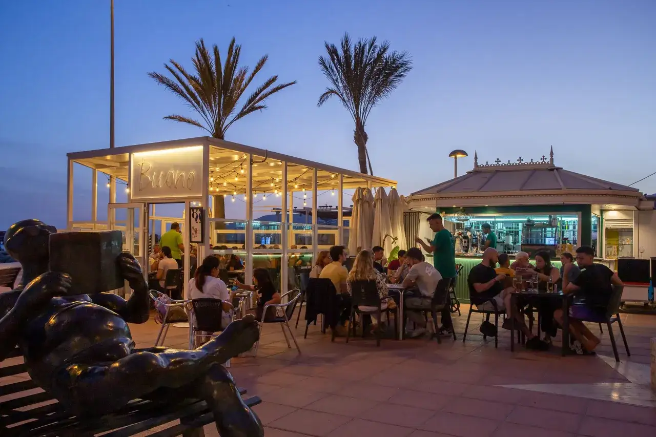 Gente cenando y bebiendo en el bar La Cala Almería al atardecer, con palmeras y luces cálidas.