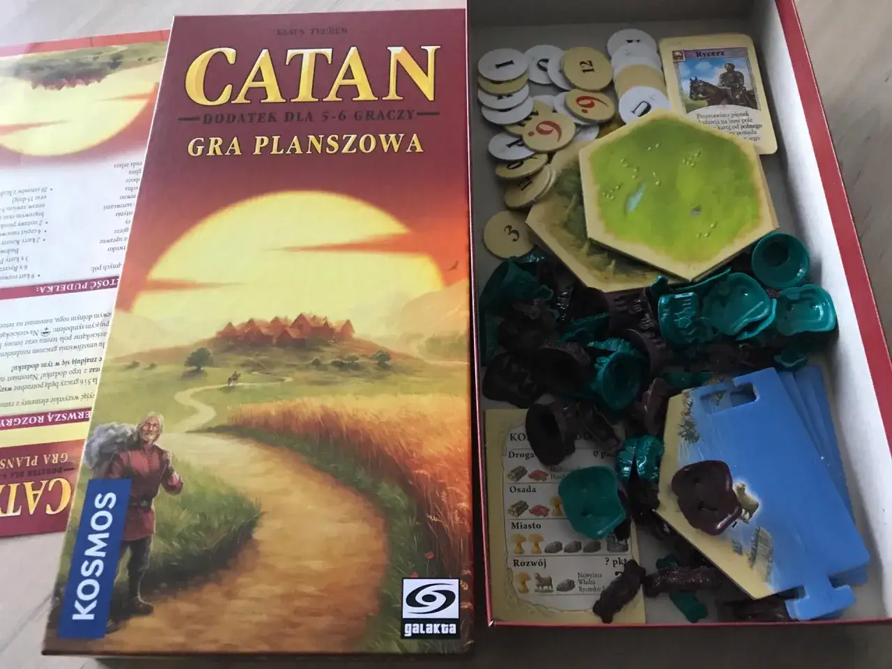 Pudełko z dodatkiem do gry Catan, zawierające elementy do gry dla 5-6 graczy. W środku widoczne są żetony, karty i pionki. Kolejność rozpakowywania element&oacute;w nie ma znaczenia.