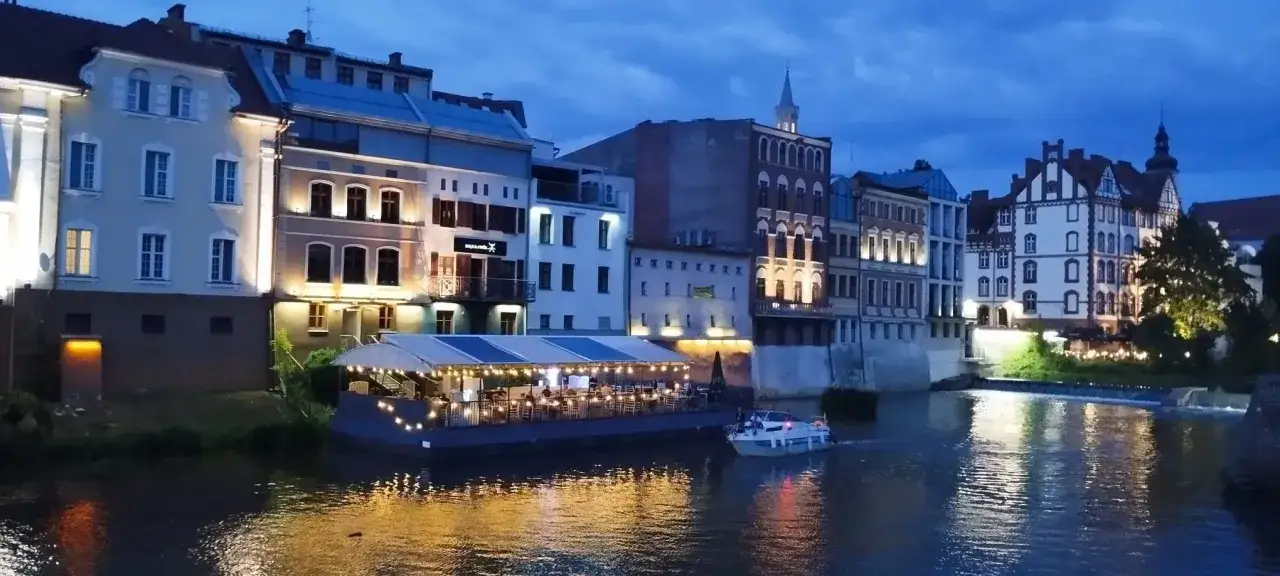 Wieczorem Odrą płynie łódka, a nad nią unosi się restauracja na wodzie. Zabytkowe kamienice tworzą malowniczy widok, niczym opolska Wenecja.