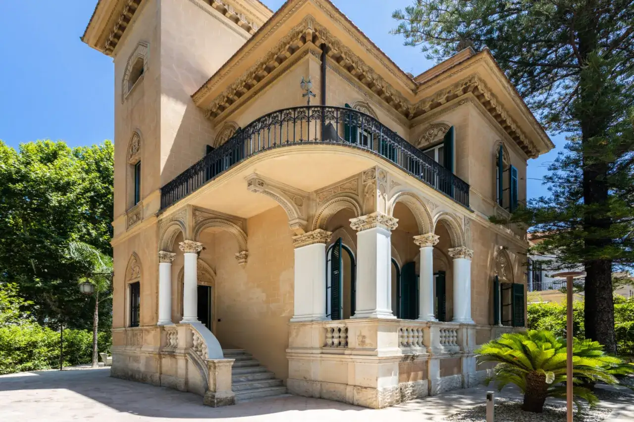 Villa in stile liberty siciliano con balcone in ferro battuto, colonne bianche e dettagli decorativi.