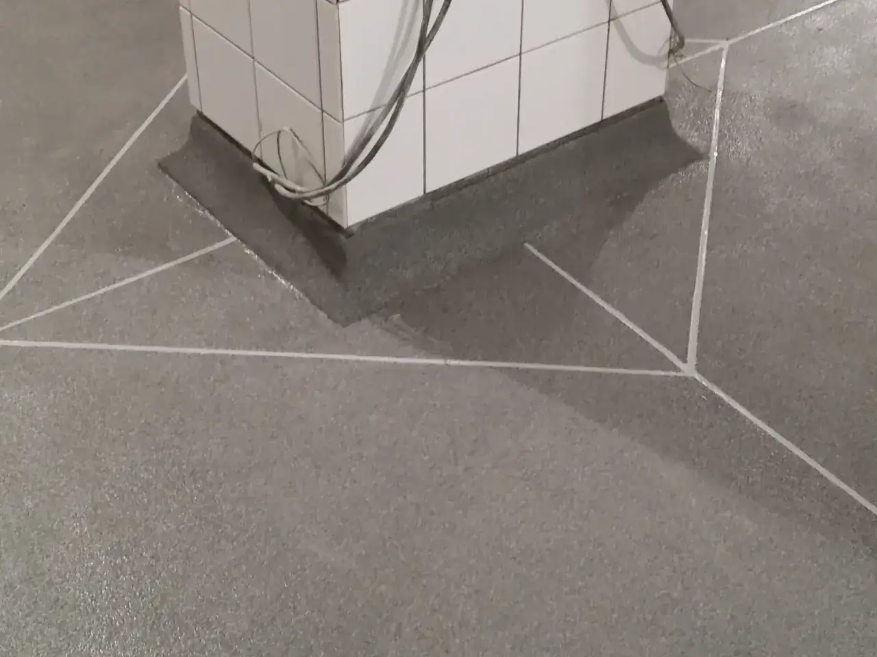 Szara posadzka betonowa z białymi liniami tworzącymi geometryczny wzór. W centrum biała, kafelkowana podstawa z kablami. Jaki beton na posadzkę? Ten wygląda na gładki i wytrzymały.