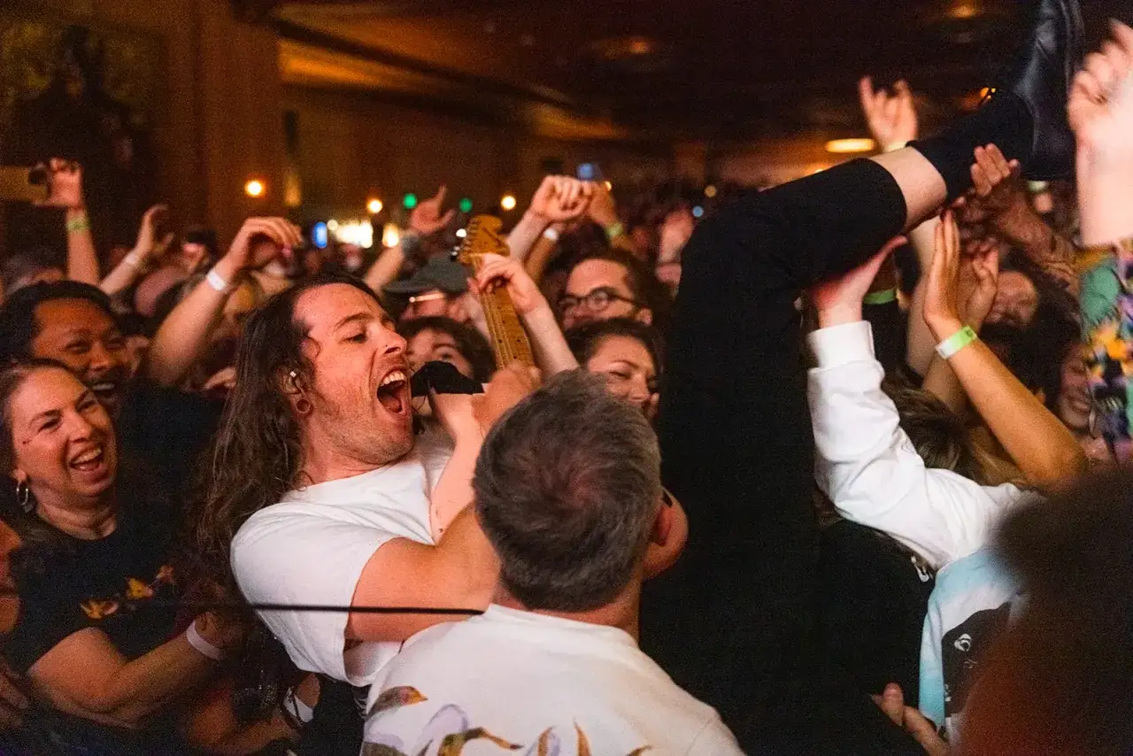 A singer with long hair belts into a mic, surrounded by a cheering crowd. Someone's legs are lifted high, a testament to the energy of the idles setlist.