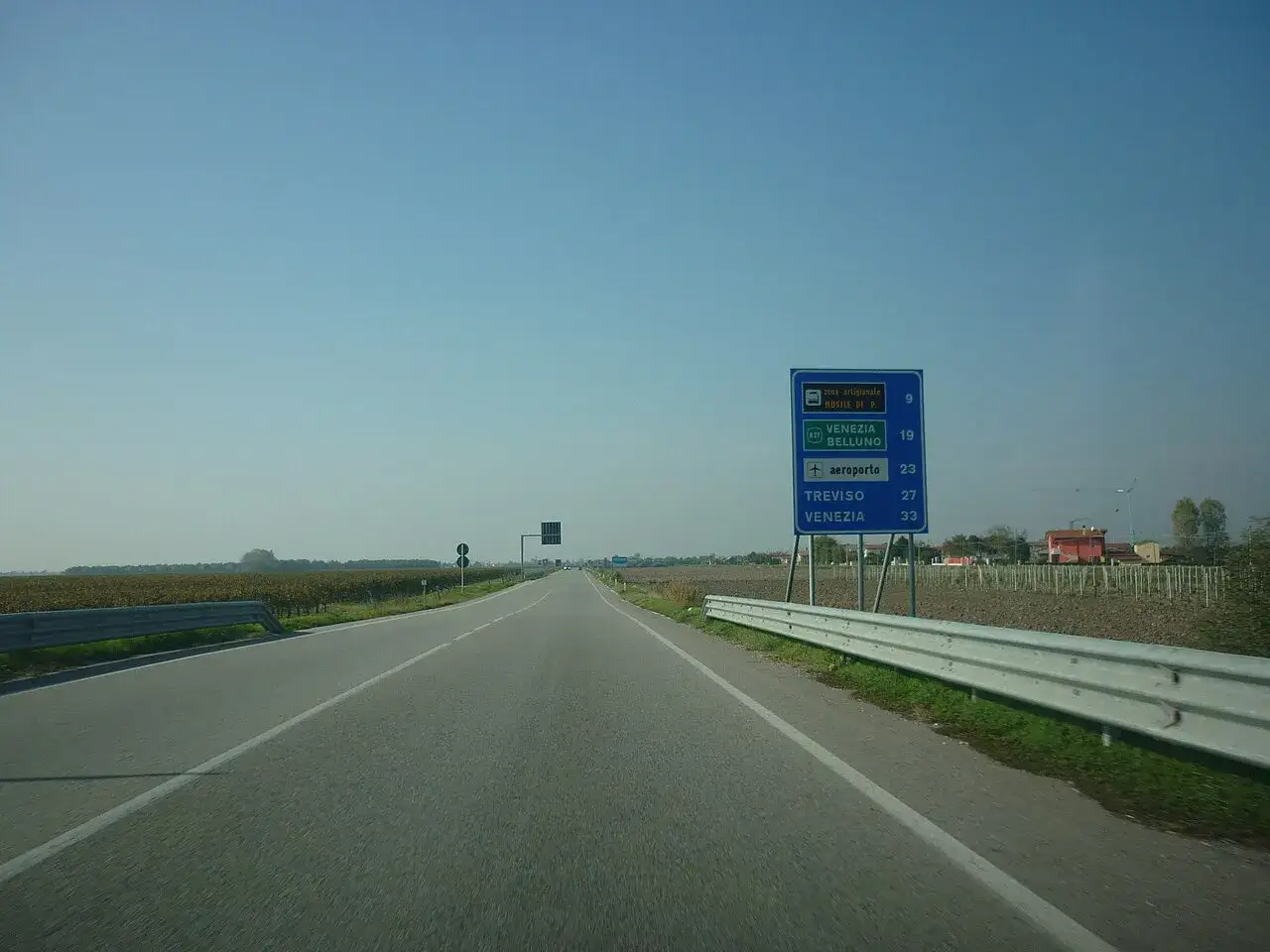 Strada extraurbana secondaria con cartello direzionale per Venezia, Belluno, Treviso e aeroporto. Campi coltivati ai lati.