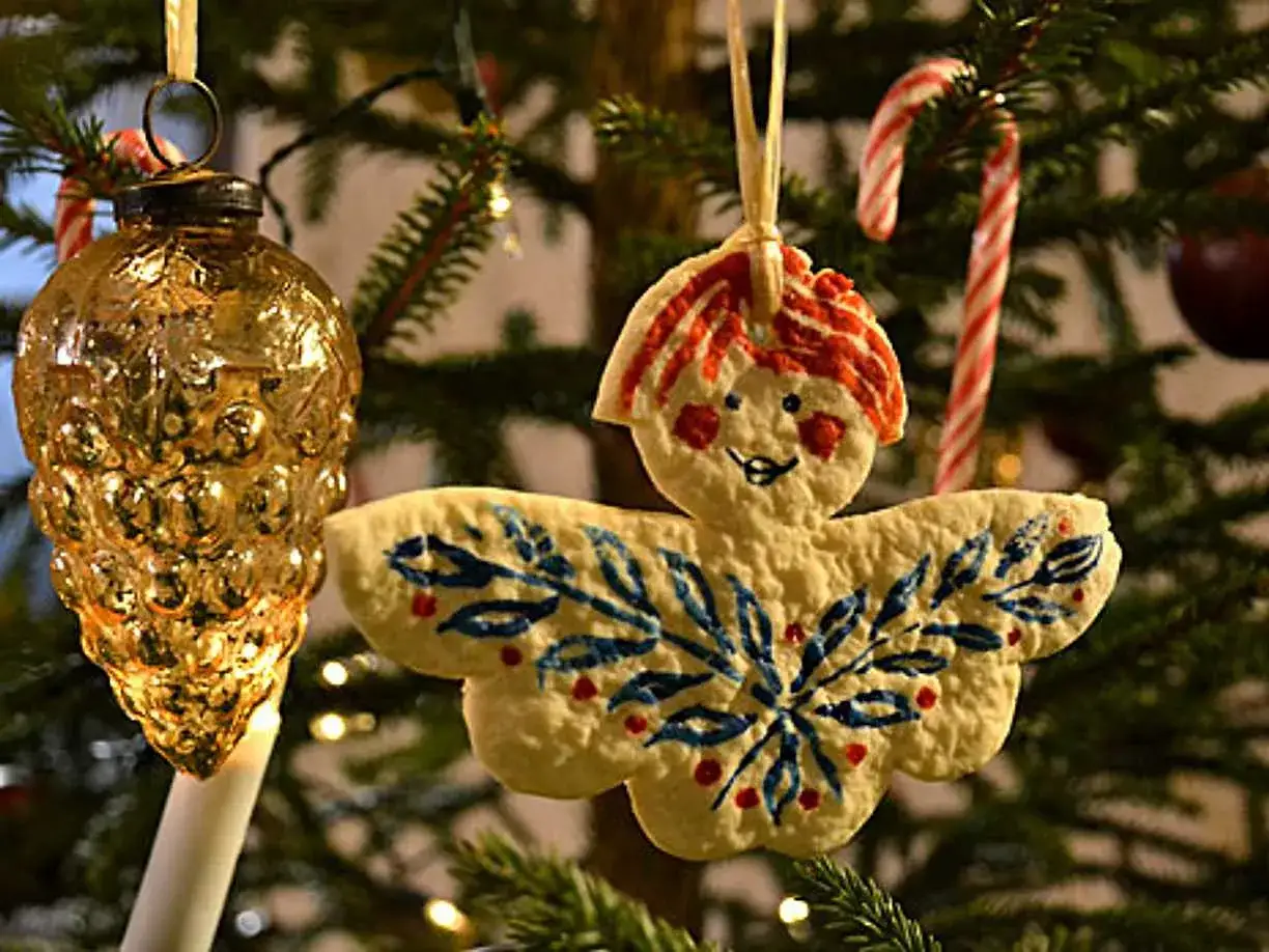 En &auml;ngel av vita pepparkakor h&auml;nger p&aring; julgranen, dekorerad med bl&aring;a blad och r&ouml;da b&auml;r. Bredvid syns en guldf&auml;rgad julgranskula.