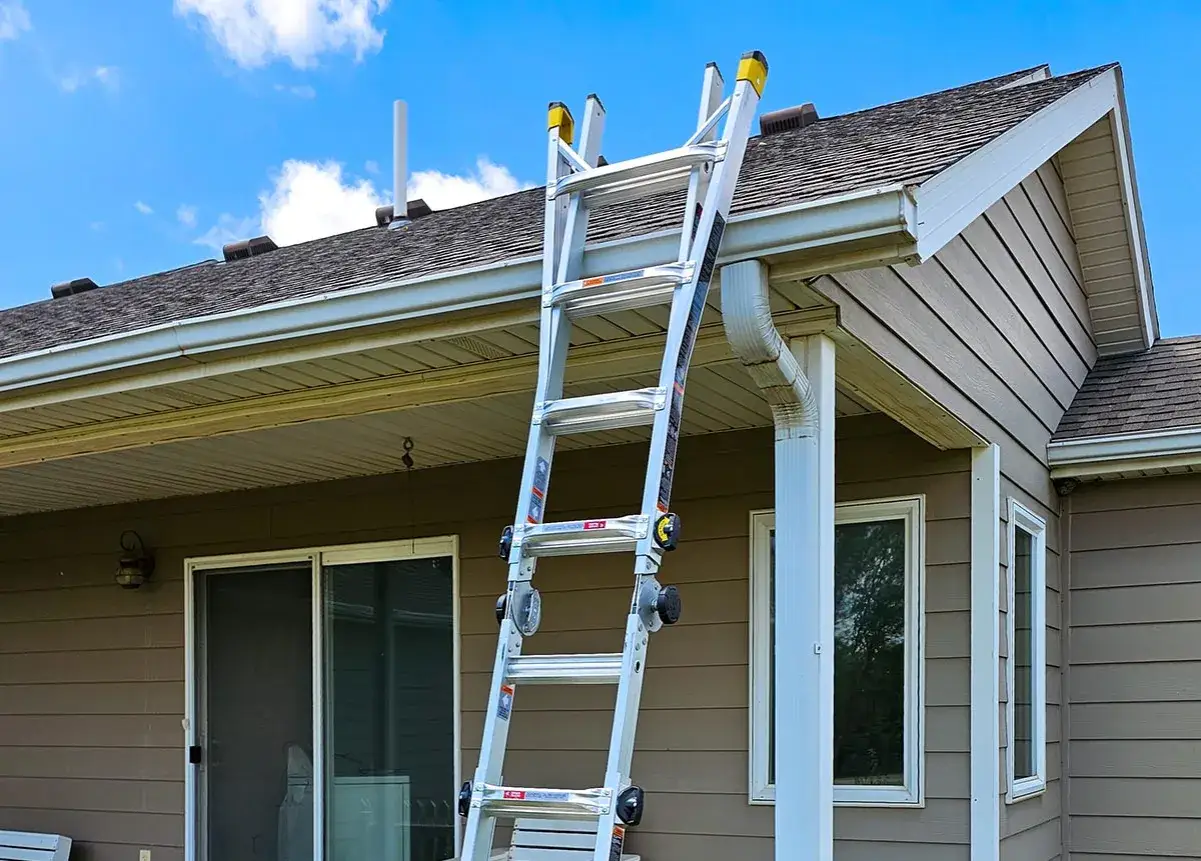 Ladder leaning against a house, ready for gutter cleaning. The image shows the roofline, gutters, and downspout.