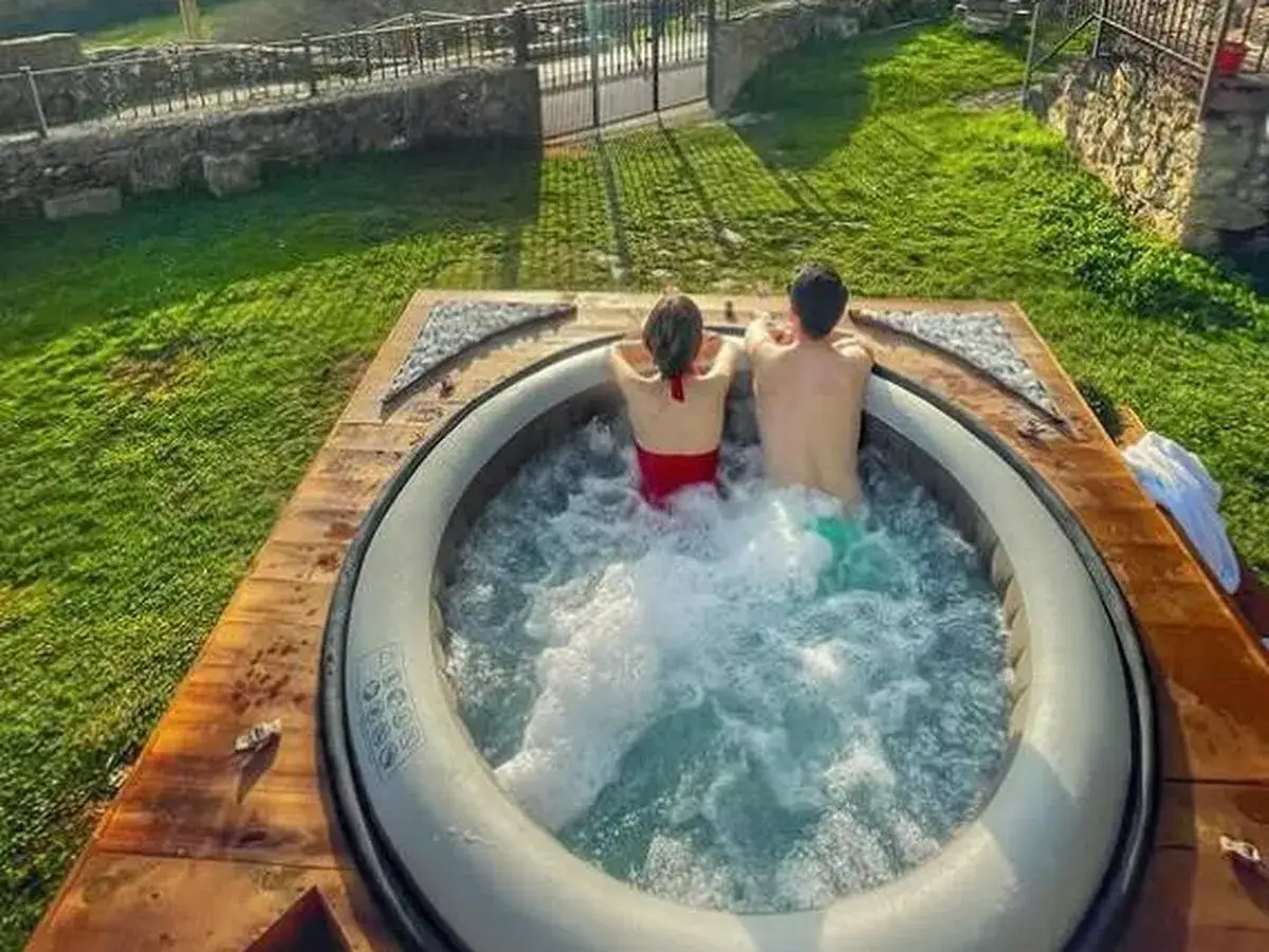 Una pareja disfruta de una relajante experiencia en un jacuzzi al aire libre, rodeados de c&eacute;sped y un entorno r&uacute;stico.