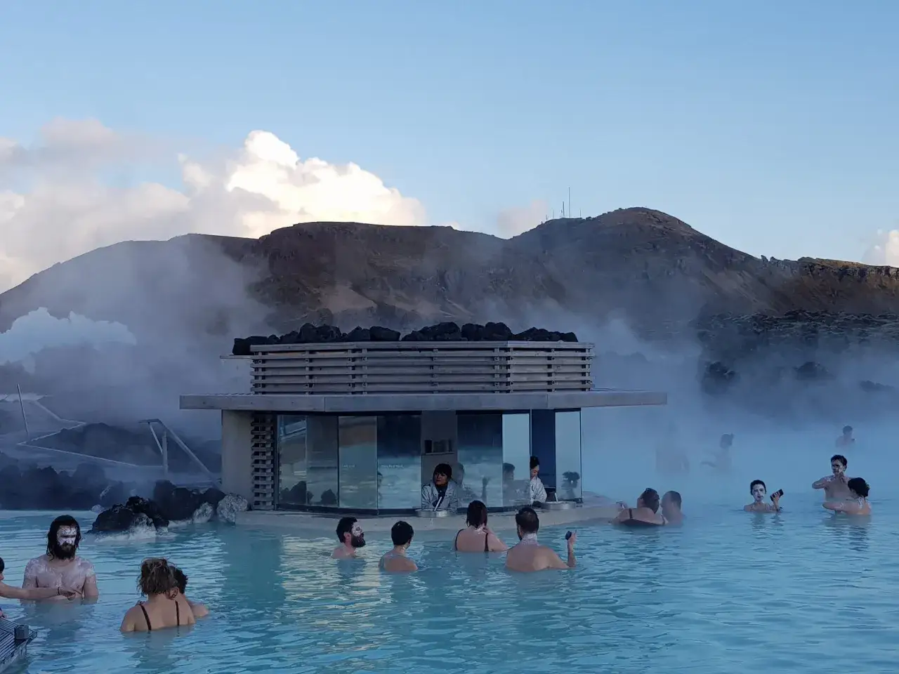Ludzie kąpią się w turkusowej wodzie Blue Lagoon na Islandii, z parą unoszącą się w tle i górskim krajobrazem.