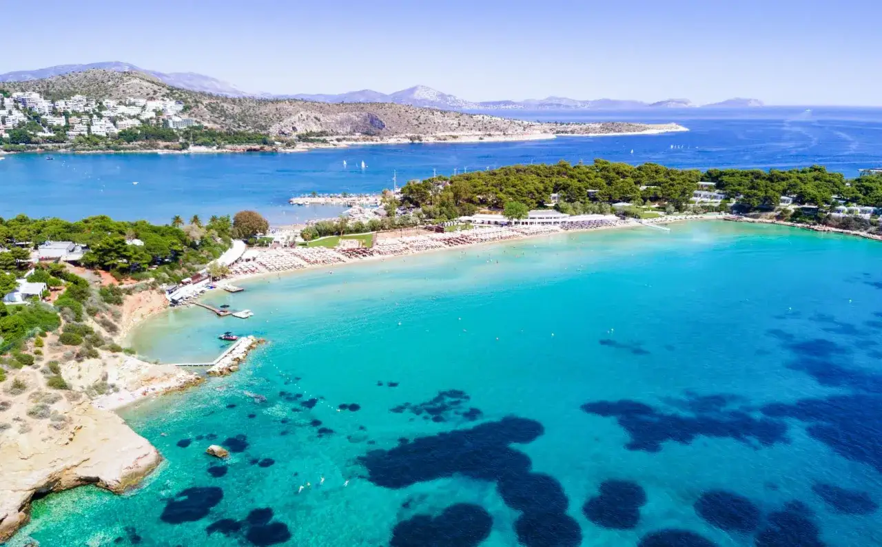 Traumhafter **Strand Athen** mit t&uuml;rkisfarbenem Wasser, Liegest&uuml;hlen und gr&uuml;nen H&uuml;geln im Hintergrund.