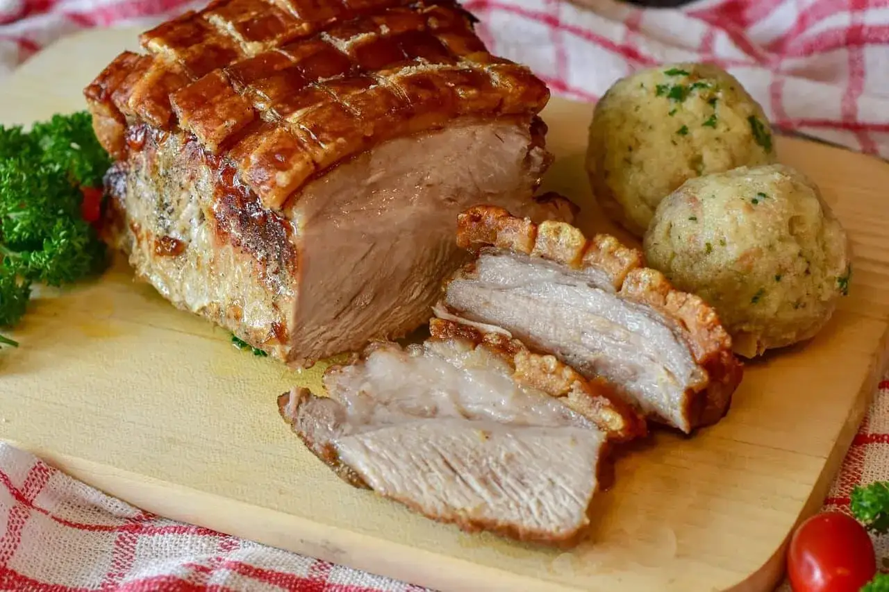 Un délicieux roti de porc avec une couenne croustillante, servi avec des boules de pain et du persil sur une planche en bois.