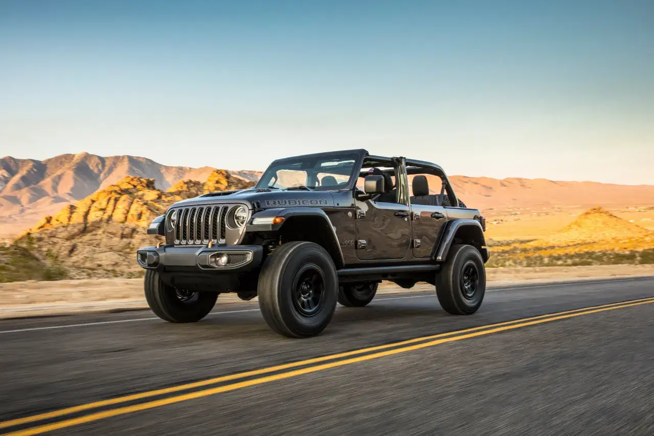 Un coche Rubicon negro avanza por una carretera des&eacute;rtica al atardecer, con monta&ntilde;as doradas de fondo.