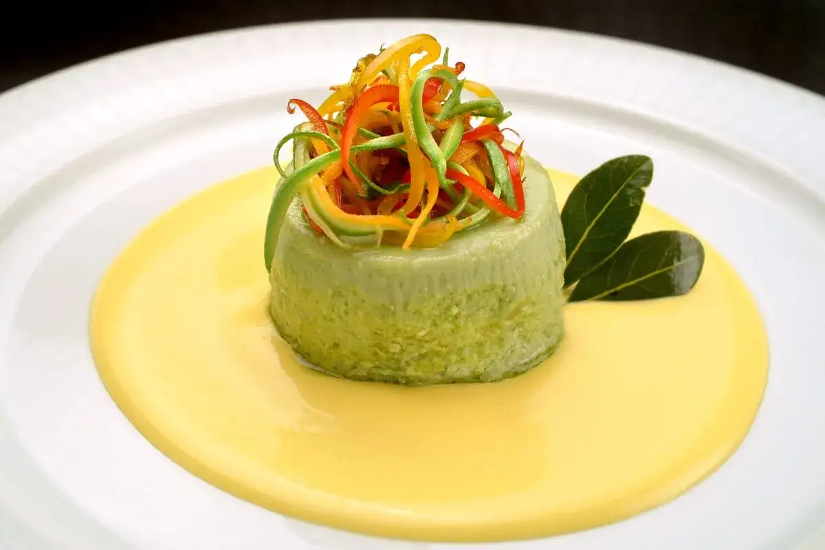 Delicato flan di carciofi su crema gialla, guarnito con julienne di peperoni colorati e foglie verdi.