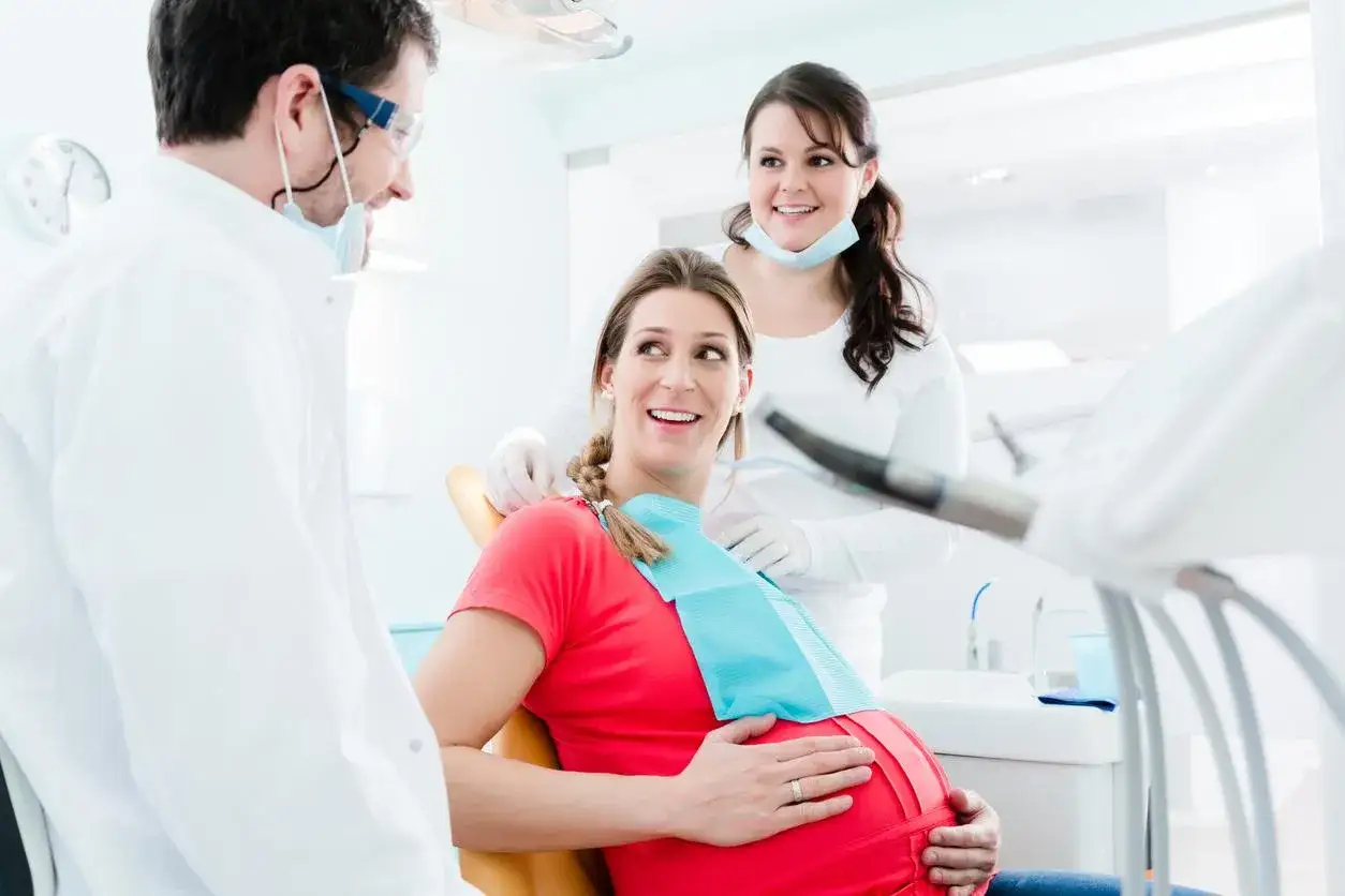 Dentista visita donna incinta con mal di denti in gravidanza. Sorridono mentre lei si tiene la pancia.
