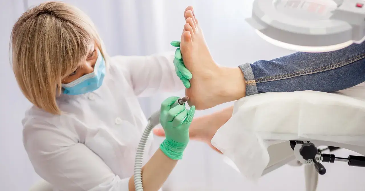 Profesjonalny pedicure co to? Kosmetyczka w maseczce i rękawiczkach wykonuje zabieg frezarką na stopie klientki.