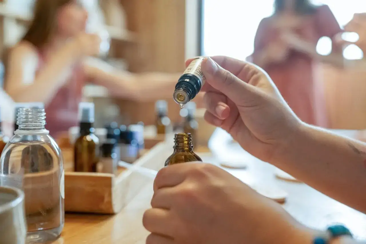 Une main verse une goutte d'huile essentielle d'un flacon dans un autre. Des flacons sont dispos&eacute;s sur une table en bois.