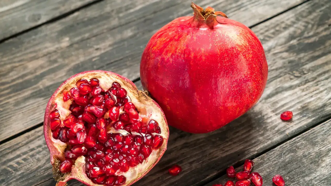 Ein ganzer und ein halber Granatapfel liegen auf Holz. Die leuchtend roten Kerne des halben Granatapfels sind sichtbar. Woher kommt der Granatapfel?