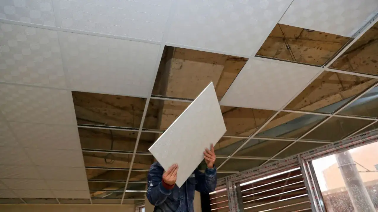 Pracownik montuje płyty do ocieplenia sufitu podwieszanego. Widać odsłonięte betonowe stropy i metalową konstrukcję.
