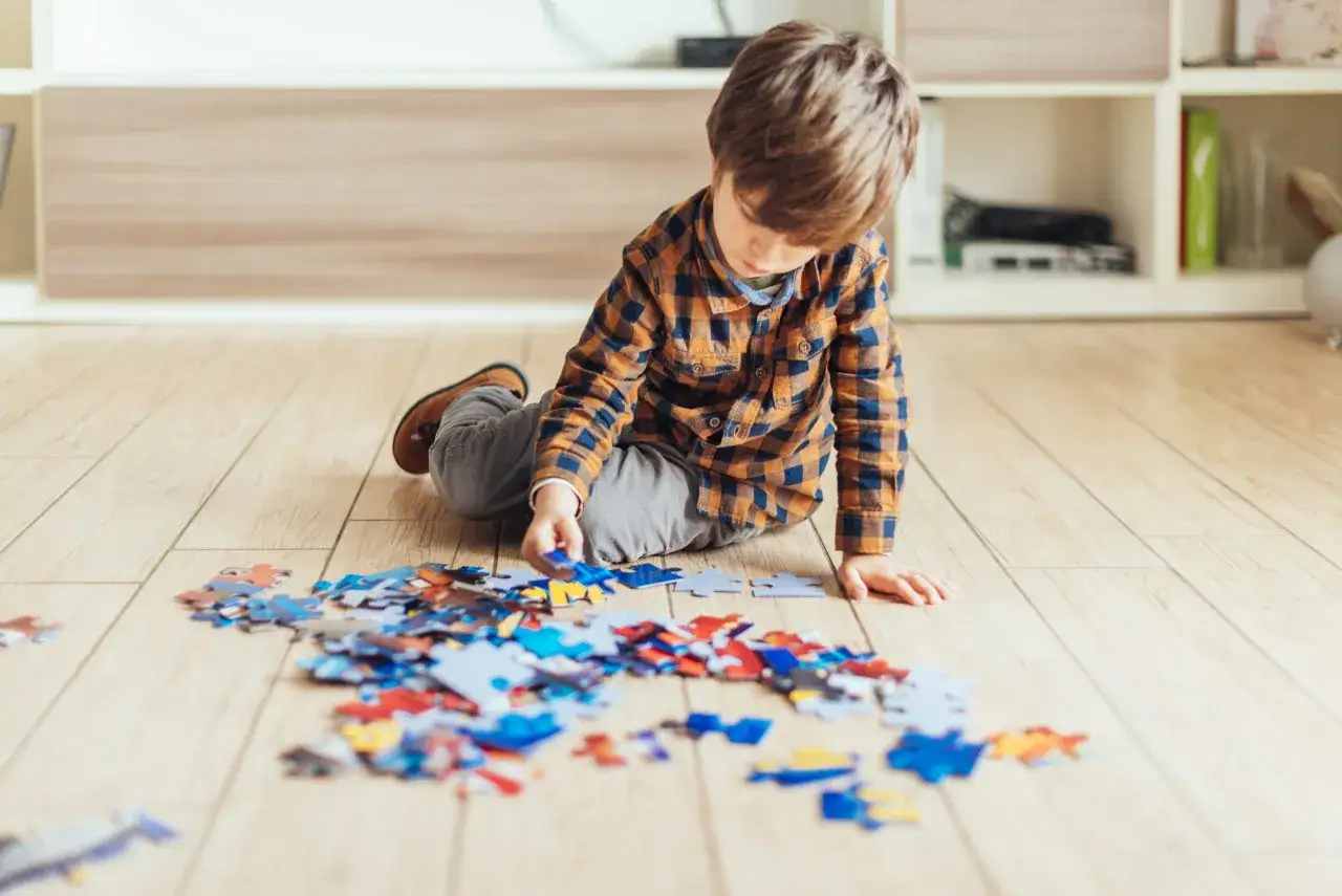 Chłopiec w kratę skupiony na układaniu puzzli na drewnianej podłodze. Kolorowe kawałki rozłożone wokół niego.