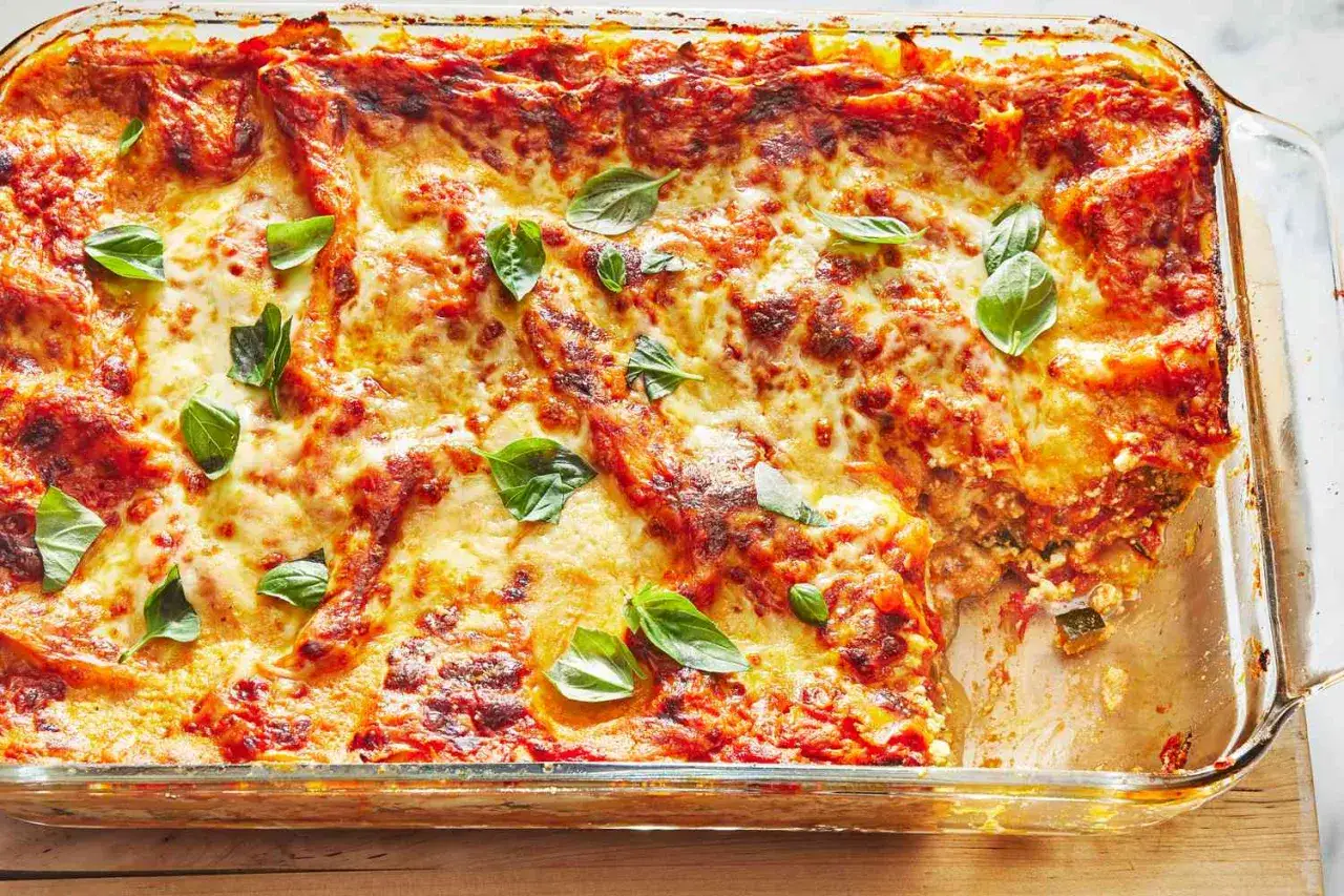 En gyllene, bubblande vegetarisk lasagne i en glasform, toppad med färsk basilika. En bit är redan uppskuren, vilket avslöjar lager av pasta, ost och grönsaker.