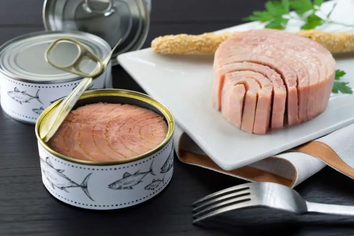 Tonno in scatola, un alimento comune ma che fa male. Pezzi di tonno in scatola e un blocco affettato su un piatto, con forchetta e grissini.