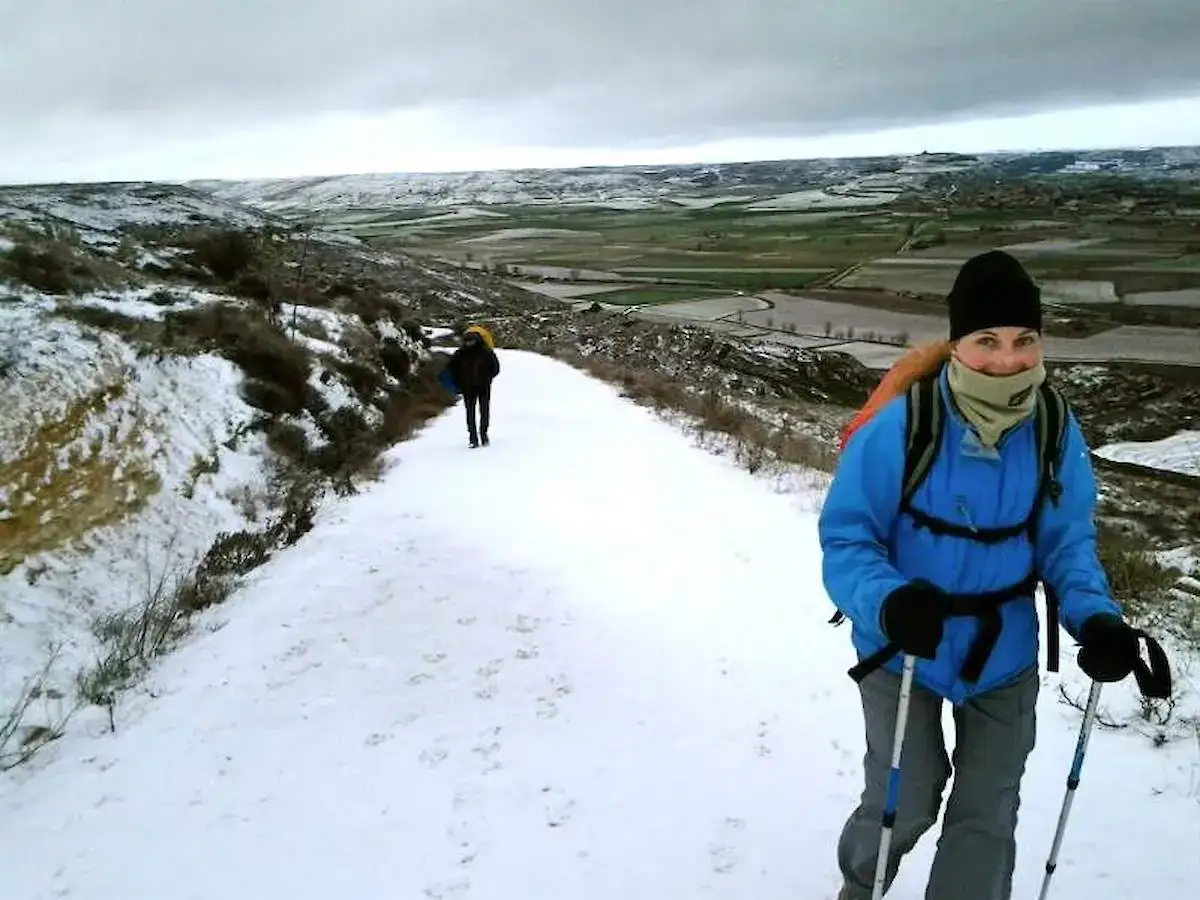 Un cammino d'inverno sotto un cielo nuvoloso. Una donna sorride mentre cammina su un sentiero innevato con bastoncini da trekking.