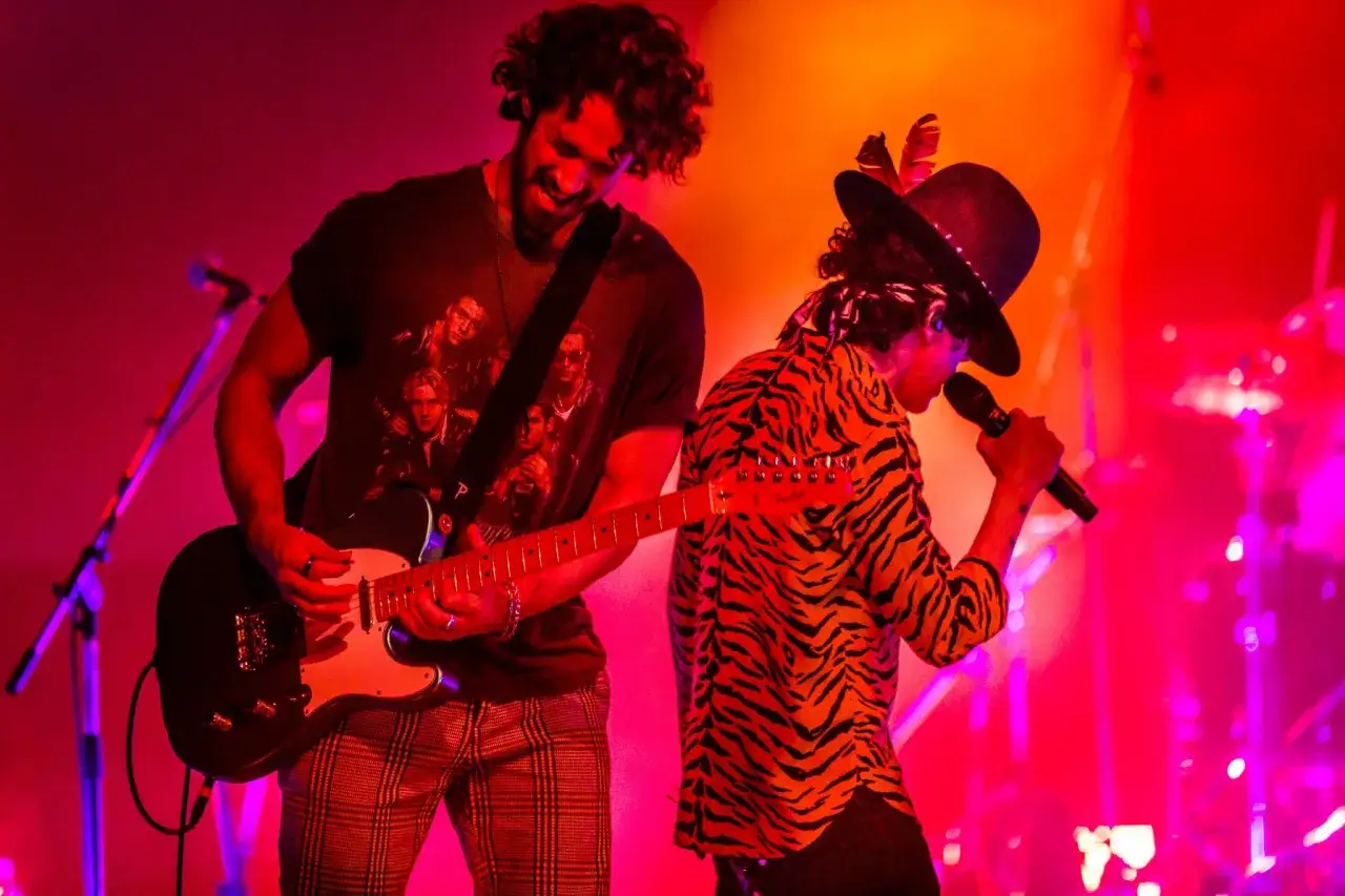 Alex Feder spielt Gitarre, w&auml;hrend ein S&auml;nger mit Hut und Tiger-Shirt auf der B&uuml;hne singt.