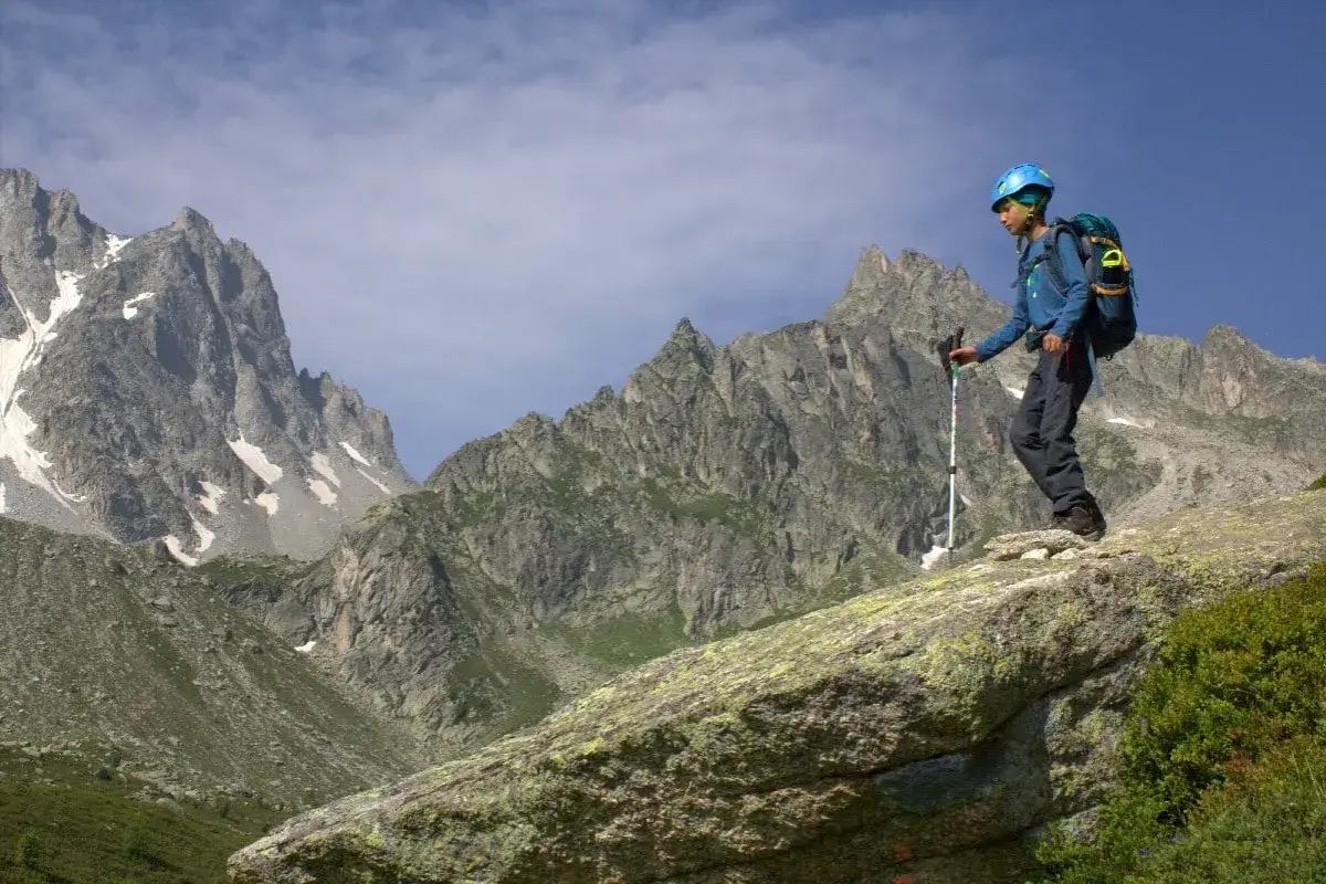 Młody turysta w kasku i z plecakiem przemierza skaliste zbocze. W tle majestatyczne szczyty Alp, część trasy Tour de Mont Blanc.
