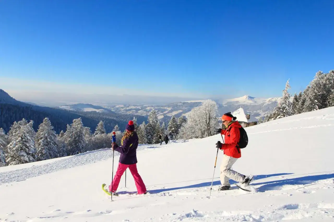 Deux personnes font de la raquette sur un sentier enneigé au Mont-Dore, profitant d'un paysage hivernal spectaculaire sous un ciel bleu éclatant.
