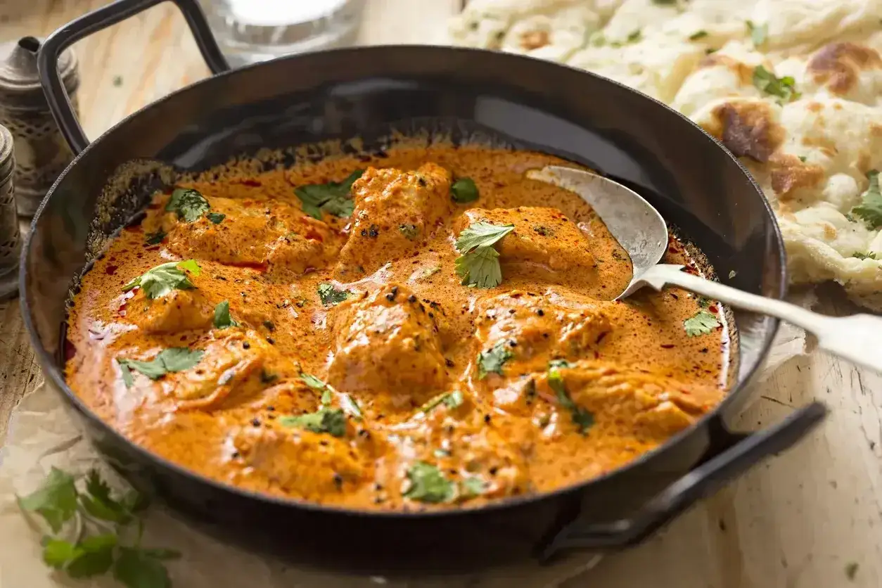 Aksamitny sos curry z kawałkami kurczaka, udekorowany świeżą kolendrą, podany w czarnej misie z łyżką. Obok pieczywo naan.