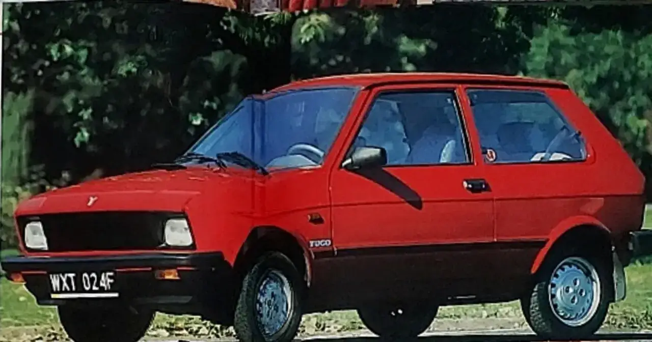 Czerwony yugo samochód z czarną atrapą chłodnicy i tablicą rejestracyjną WXT 024F, zaparkowany na tle zieleni.