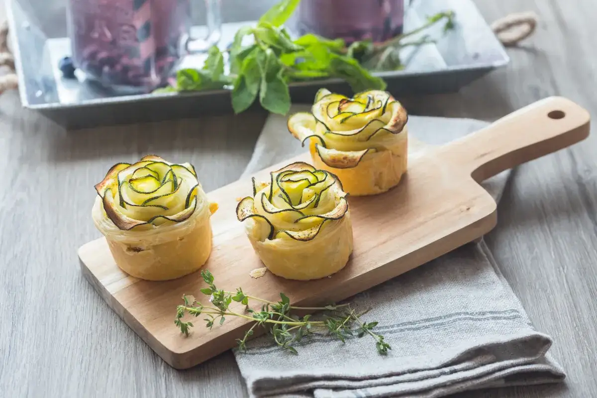 Trois tartelettes dorées ornées de fines lamelles de fleur de courgette disposées en rosace, sur une planche en bois.