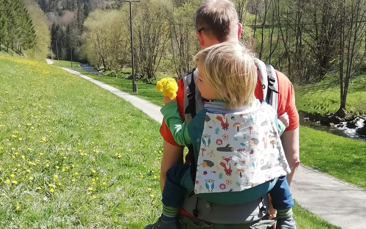 Ein Vater genießt das Wandern mit Baby auf einem sonnigen Weg entlang eines Baches. Das Kind hält eine Löwenzahn-Wolke.