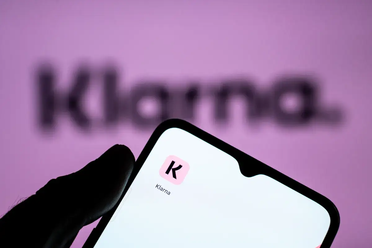 Dłoń trzyma smartfon z ikoną aplikacji Klarna. W tle logo Klarna. Zastanawiasz się klarna co to? To płatności odroczone.