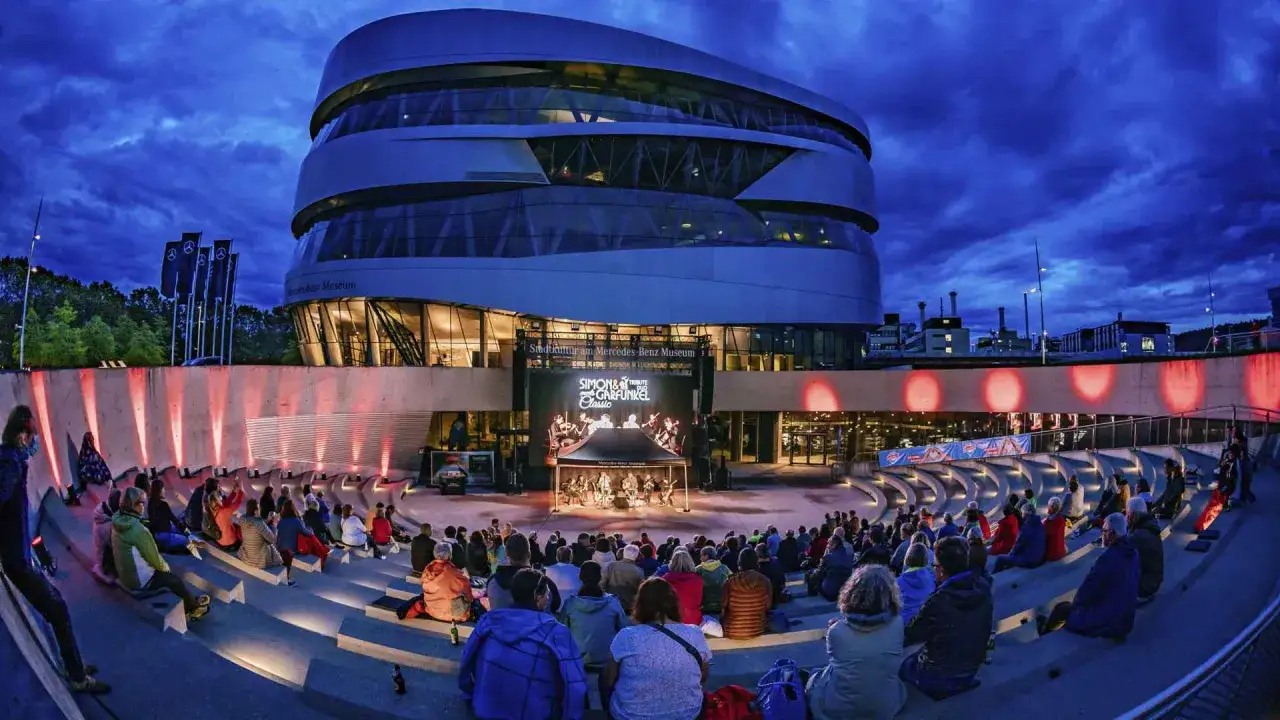 Ein Konzert im **Mercedes-Benz Museum Open Air**. Menschen sitzen auf einer Tribüne vor der Bühne, die vom Museum beleuchtet wird.