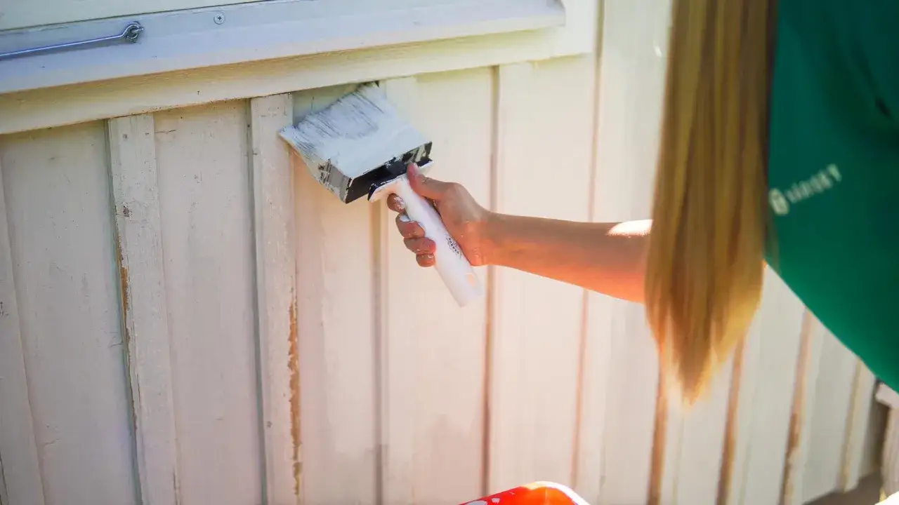 En person med långt blont hår målar en vit träfasad med en bred pensel. Det är dags att måla av huset för sommaren.