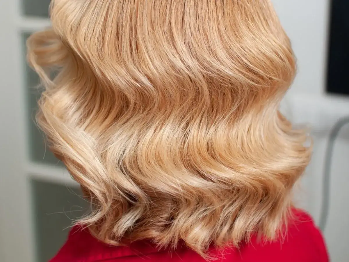 Blond włosy z delikatnymi falami, ułożone w stylizacji retro. Proste cięcie (blunt cut) nadaje im objętości i elegancji.