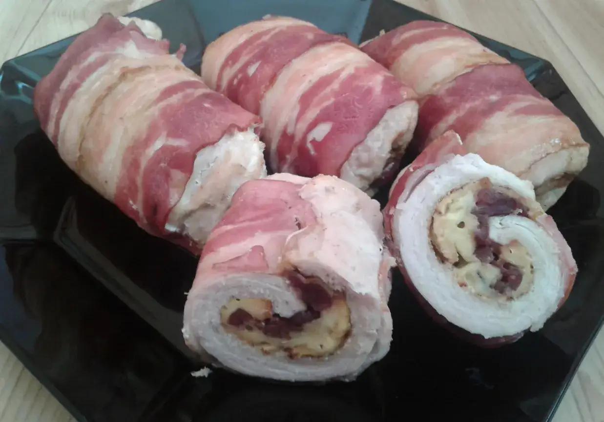 Roladki z indyka z boczkiem, gotowe do pieczenia, prezentują się apetycznie na czarnym talerzu. Widać przekrój jednej z nich, ukazujący farsz.