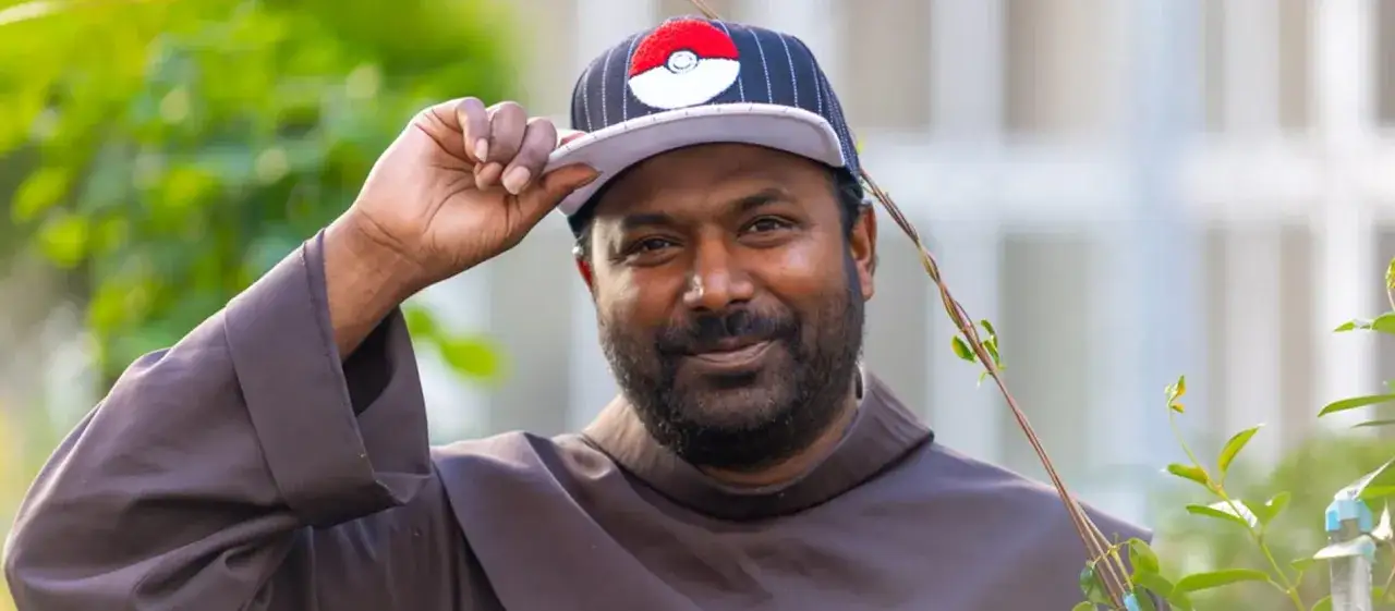Pater Sandesh Manuel, ein Mann mit Bart, trägt eine Kappe mit Pokémon-Logo und eine braune Robe.