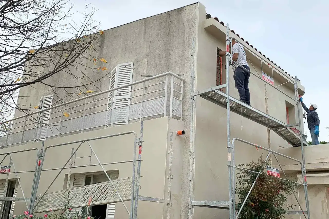 Travaux d'application d'un enduit isolant extérieur sur une maison. Des ouvriers sur échafaudage appliquent le revêtement.