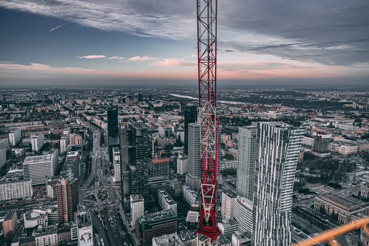 Widok z lotu ptaka na Warszawę z dominującą czerwoną wieżą budowlaną. Widać nowoczesne wieżowce, w tym Warsaw Towers, oraz rozległe miasto.