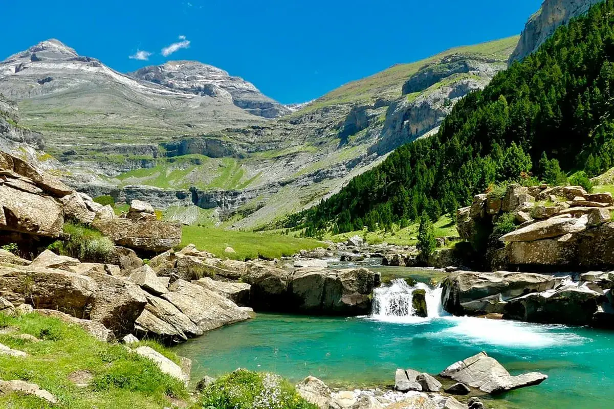 Un río turquesa serpentea entre rocas y prados verdes, con cascadas y montañas imponentes al fondo. Un paisaje idílico que recuerda a los ibones de Anayet.