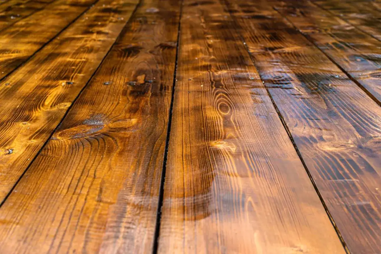 Tavolato di legno scuro, lucido e bagnato, con venature evidenti. La patina riflette la luce, creando un effetto quasi specchiato.