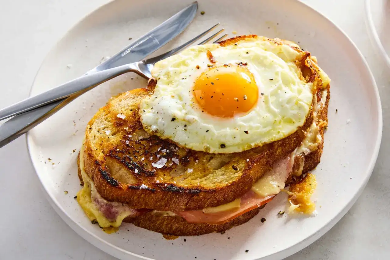 En gyllene croque madame med stekt ägg, ost och skinka på rostat bröd, serverad på en tallrik med bestick.