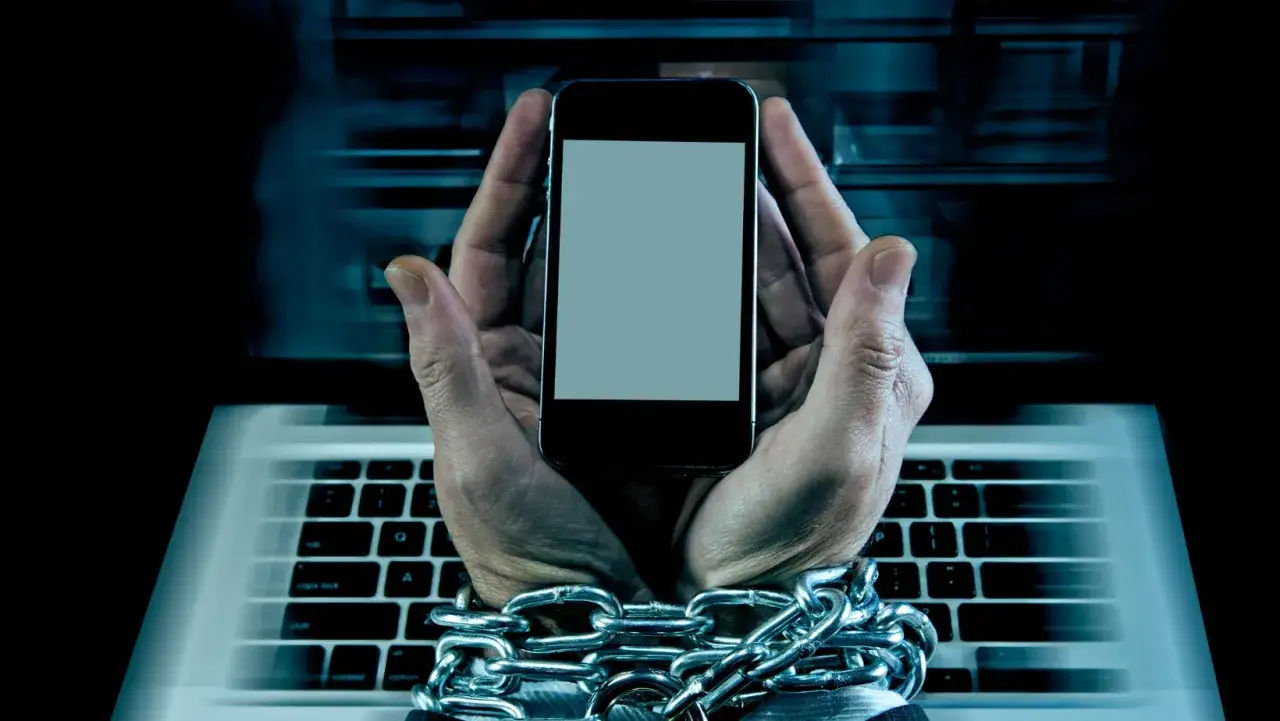 Uwięzione dłonie trzymają smartfon na tle klawiatury laptopa. Ciężkie łańcuchy symbolizują uzależnienie, być może erotomanię internetową.