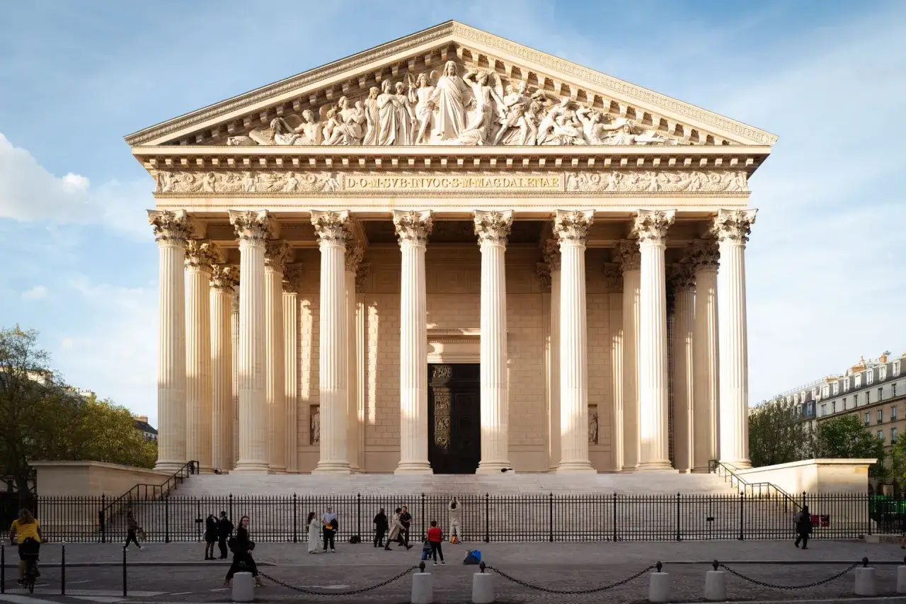 La Madeleine, tambi&eacute;n conocida como Saint Sulpice, se alza majestuosa con sus columnas corintias. Gente pasea frente a la imponente fachada.