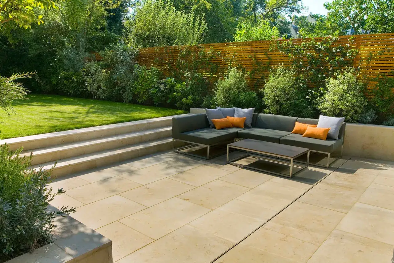 Terraza moderna con sof&aacute; y mesa de centro, escalones de piedra y un muro de contenci&oacute;n que delimita el jard&iacute;n verde.