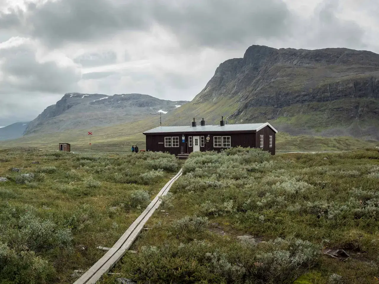 En mörk fjällstuga vid Kungsleden, omgiven av kalfjäll och låg vegetation. En träspång leder fram till stugan.