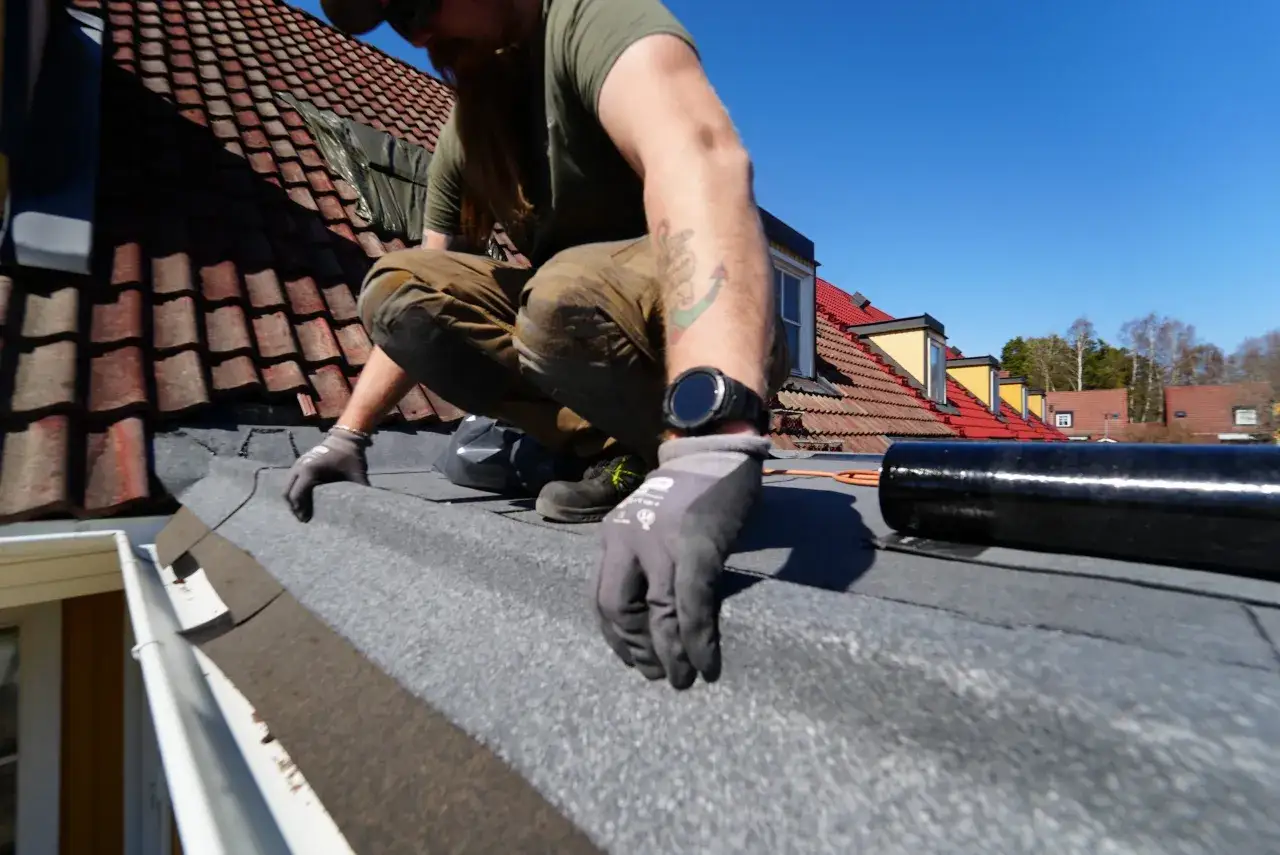 Man med skägg och tatueringar arbetar på ett tak, lägger papp. Solen skiner och himlen är blå.