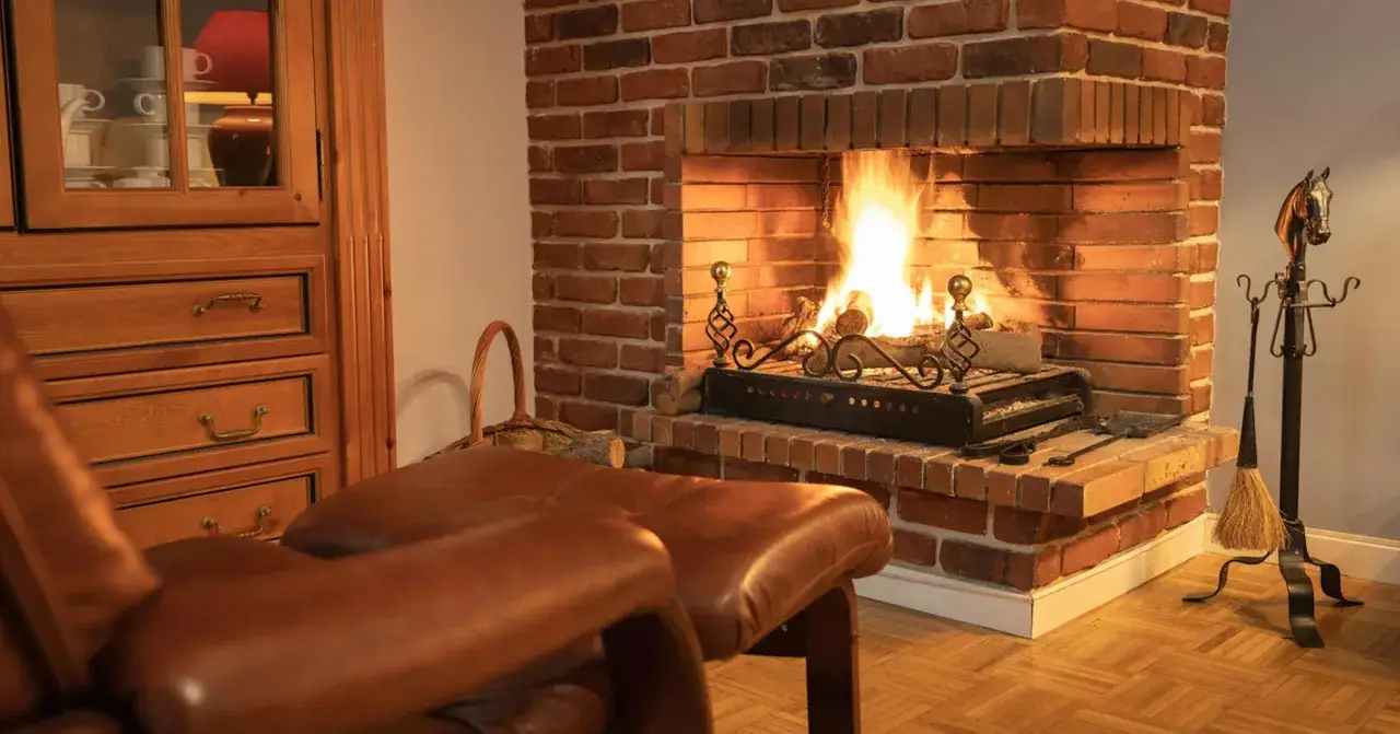 Un po&ecirc;le moderne est install&eacute; dans une ancienne chemin&eacute;e en briques, cr&eacute;ant une ambiance chaleureuse. Un fauteuil en cuir marron et une armoire en bois compl&egrave;tent la sc&egrave;ne.
