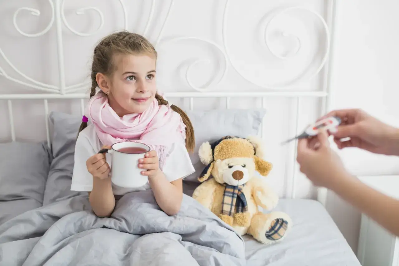 Chora dziewczynka w łóżku pije herbatę, obok siedzi miś. Mama mierzy jej temperaturę. Warto pamiętać o probiotyk dla dzieci.
