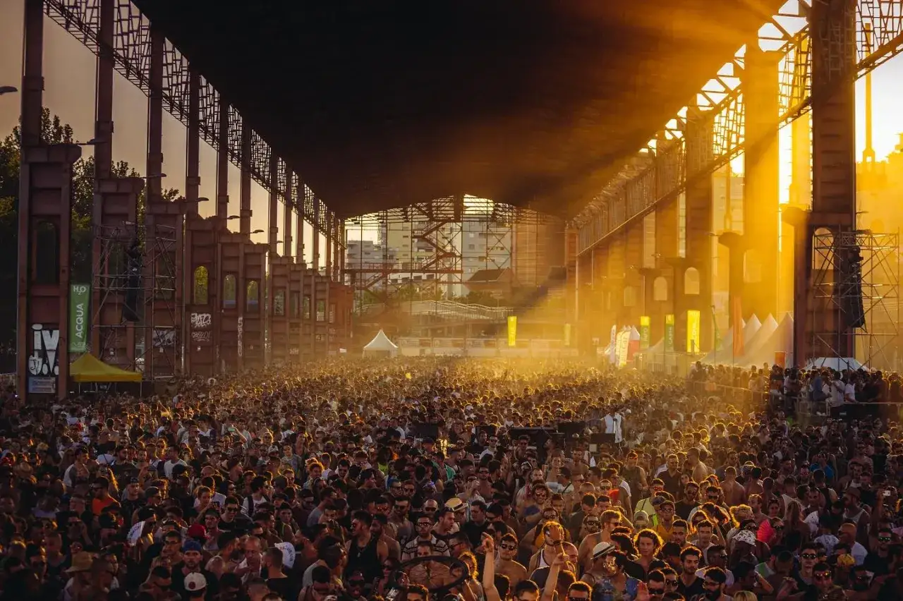 Folla immensa sotto un tetto industriale, illuminata da raggi dorati. Un concerto di gruppi musicali piemontesi in un'atmosfera magica.